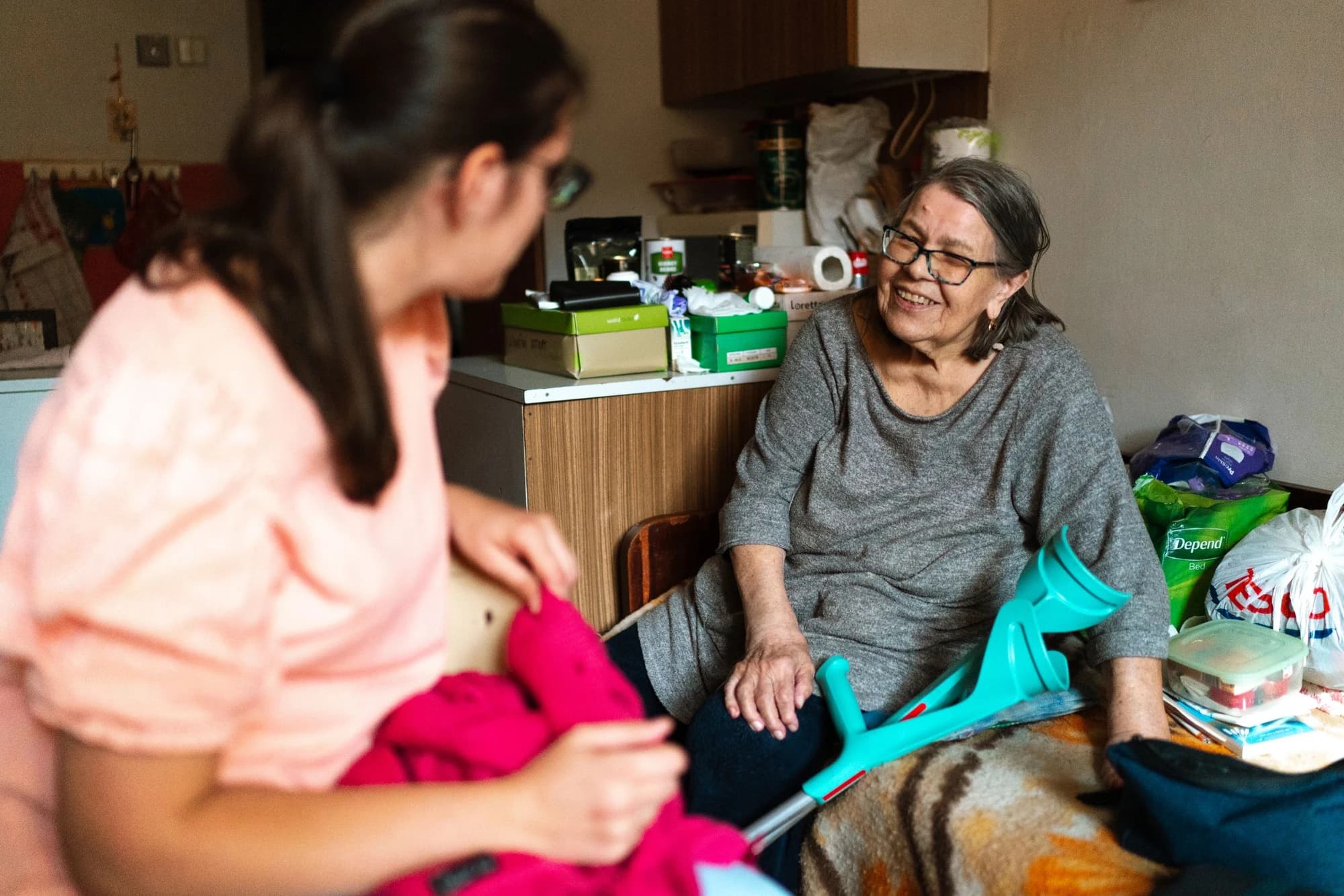 A care worker supports an older woman with a mobility aid indoors.