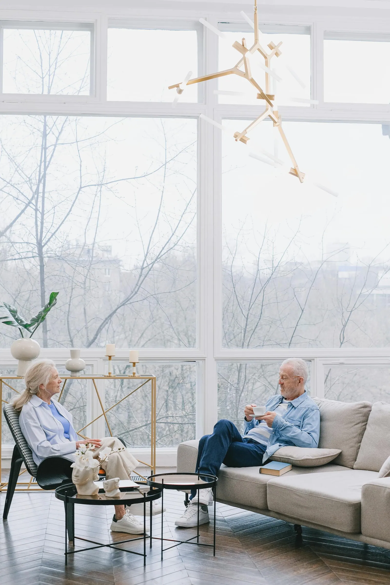 An older couple share coffee and conversation by a bright window at home.